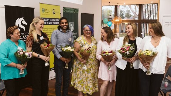 A group of six females and one male adult standing in a line smiling and each holding a bunch of flowers.